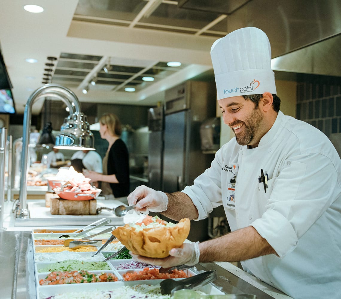 A TouchPoint chef skillfully prepares a taco bowl with fresh ingredients in a bustling kitchen.